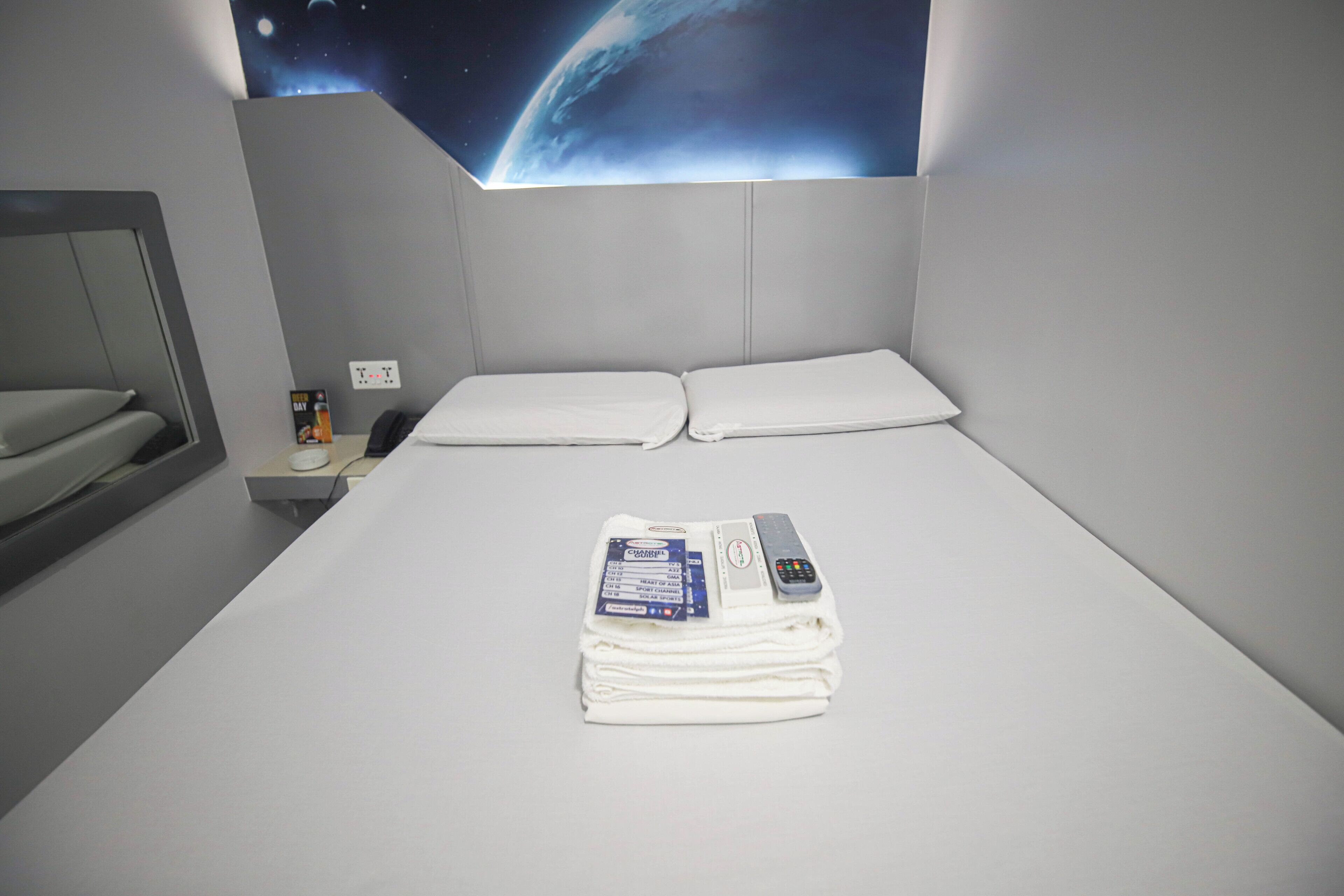 Una cama bien hecha con sábanas blancas, un control remoto y una toalla doblada en una habitación de hotel con pared gris y un mural con temática espacial.