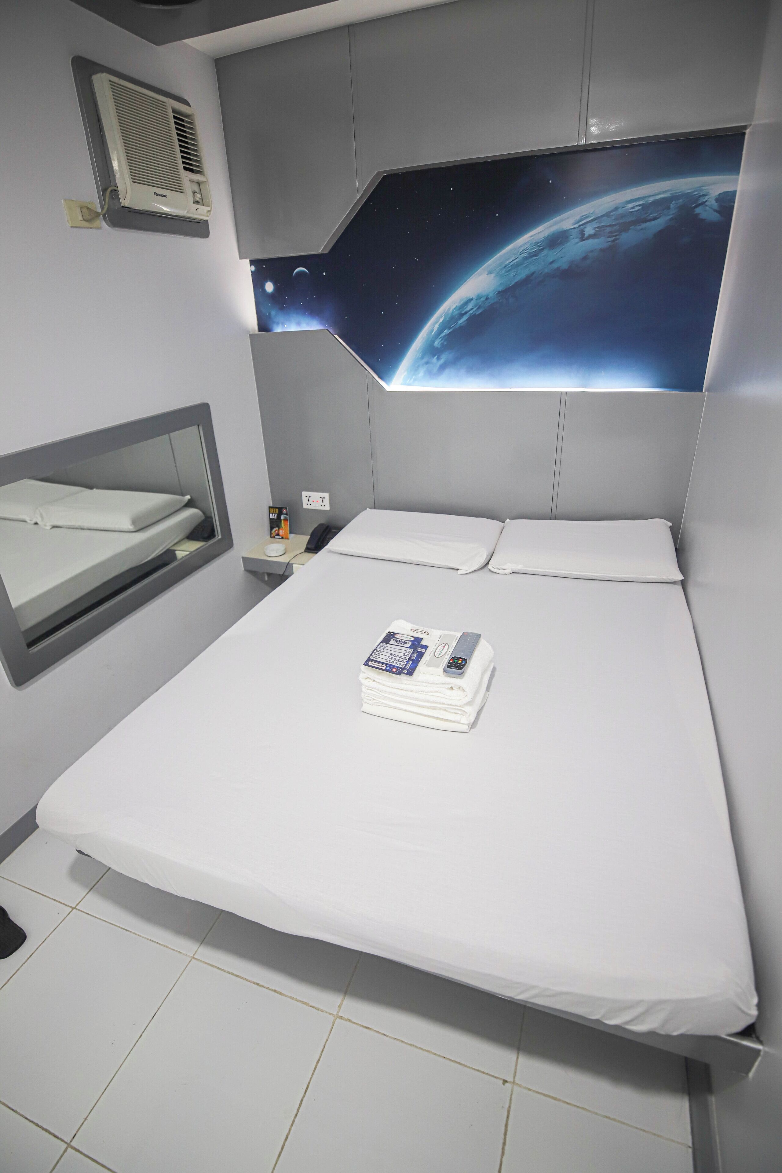 A modern hotel room with a bed, a nightstand, a mirror, a wall-mounted air conditioner, and a space-themed wall decoration.