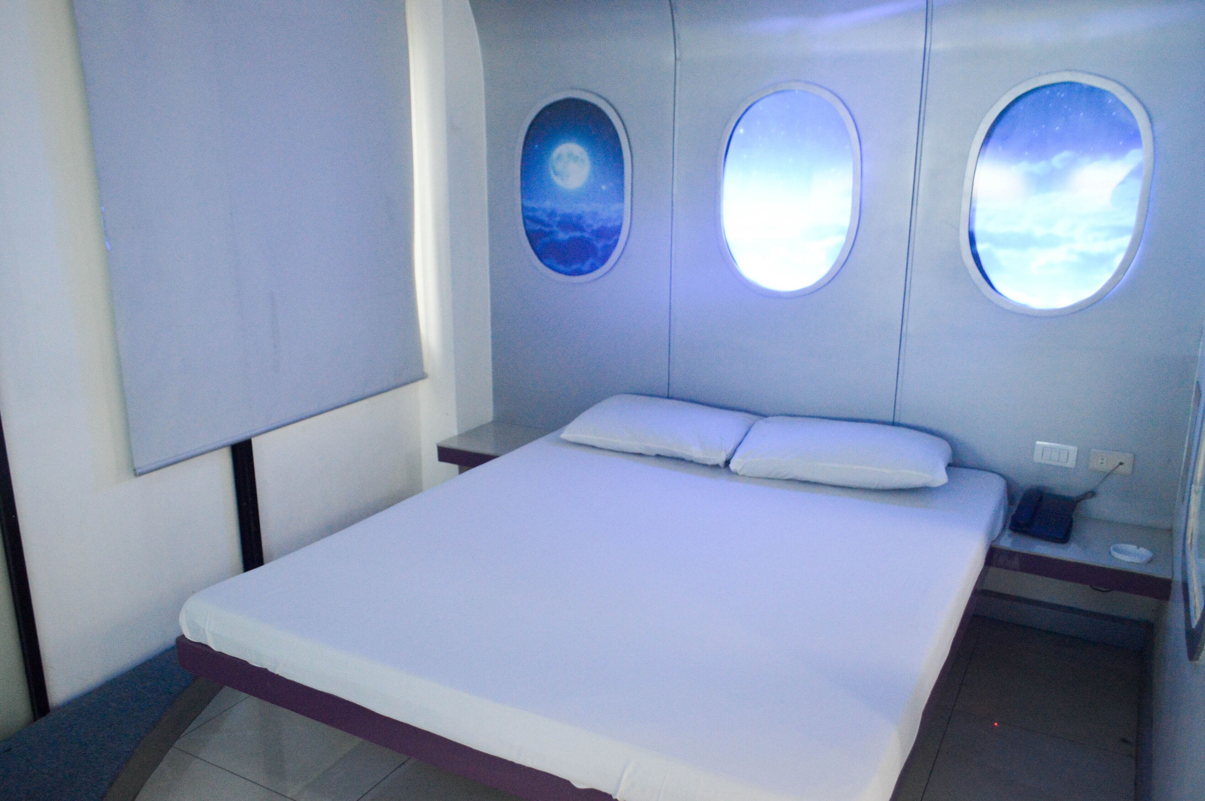A bedroom with a bed, two windows with night sky views, a bedside table, and a wall-mounted TV.