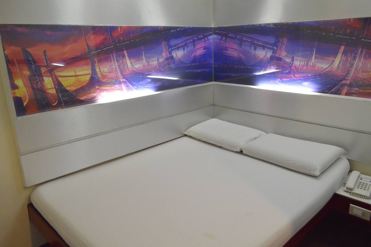 Un lit simple avec une literie blanche, un téléphone sur la table de chevet et une œuvre d’art fixée au mur représentant un paysage urbain futuriste.