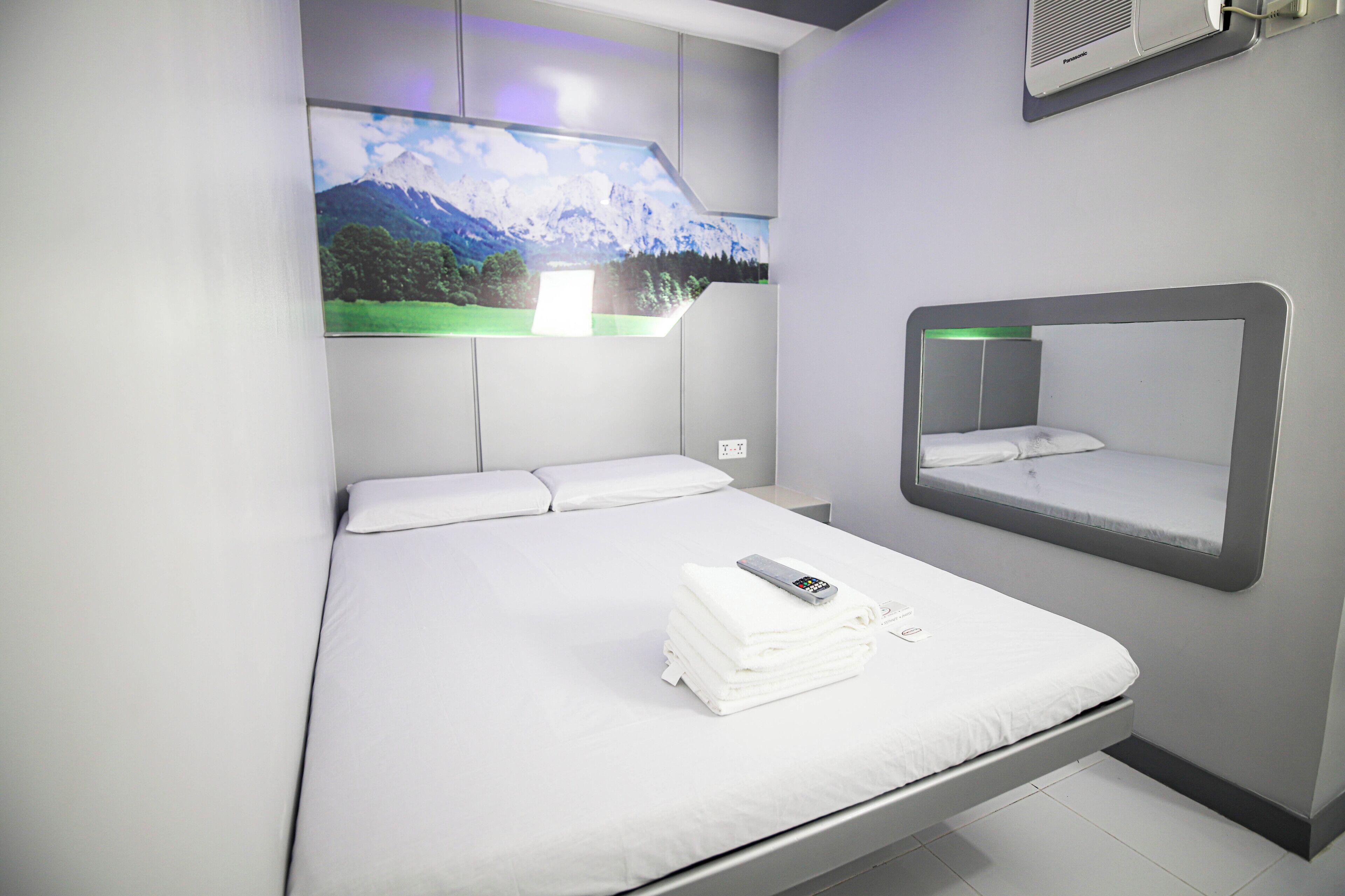 A modern hotel room with a bed, a mounted TV displaying a scenic landscape, a bedside mirror, and a remote control on the bed.