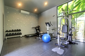 A gym with exercise equipment, a wall-mounted dumbbell rack, and a large window with a view of greenery. - Almali Villa Mali (Rawai)