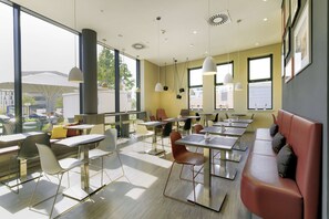 Un café moderno con mesas y sillas dispuestas en un espacio amplio, ventanas grandes que ofrecen vistas al exterior e iluminación colgante.