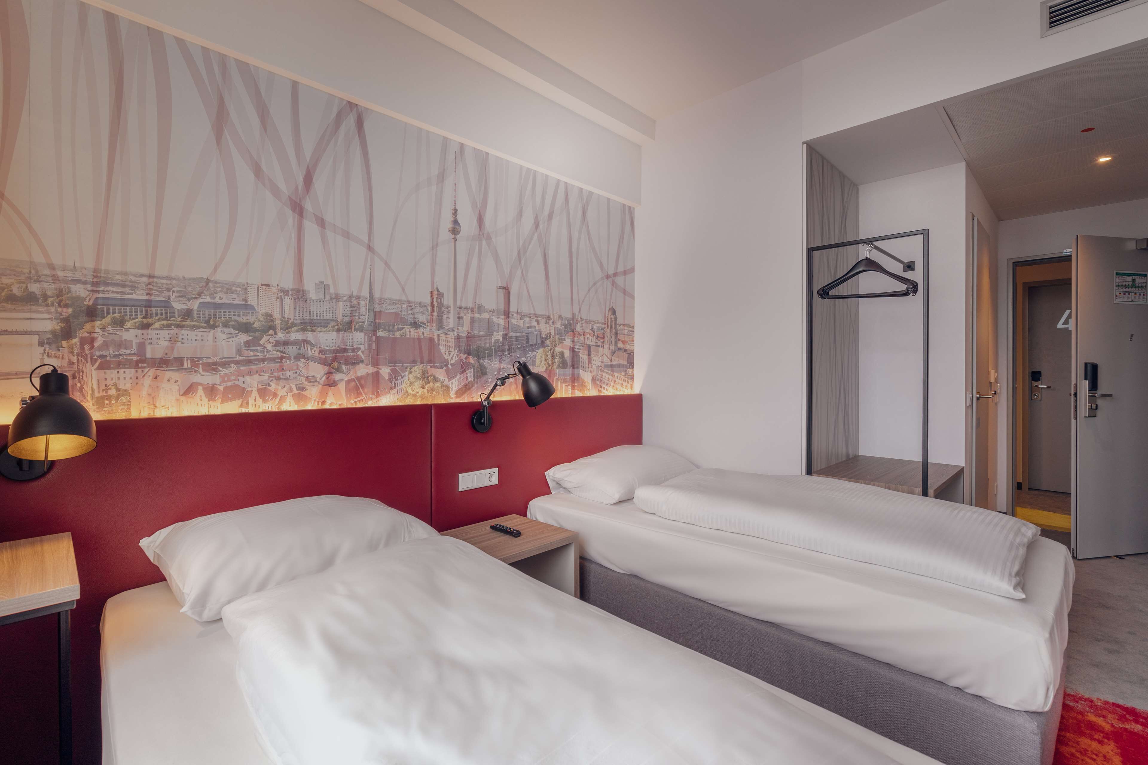 Une chambre d’hôtel avec deux lits, un mur d’accent rouge, une fresque représentant un paysage urbain, un porte-manteau suspendu et une porte donnant sur une salle de bain.