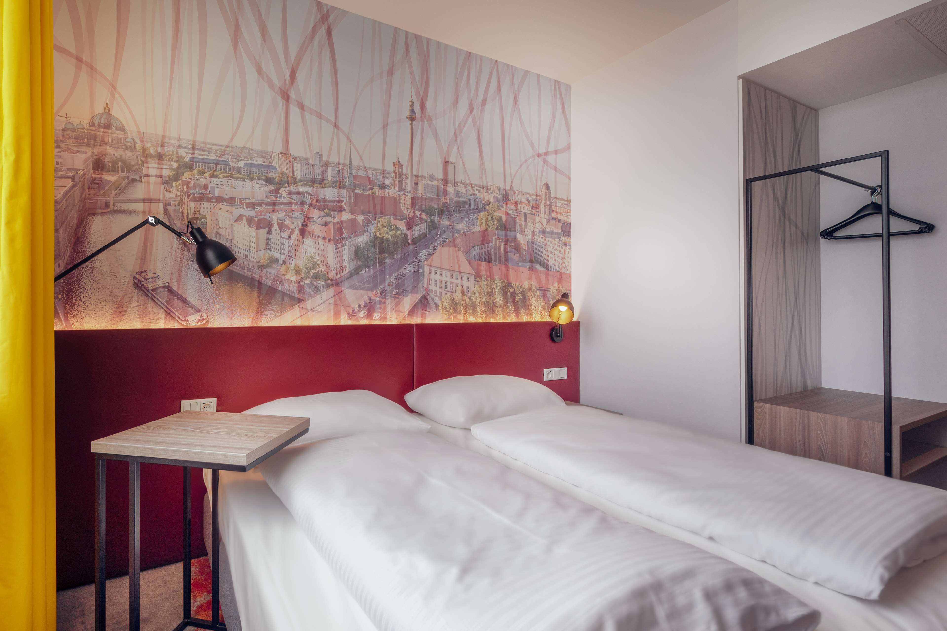 Habitació d'hotel amb dos llits, un panell cefalòtic vermell, un quadre de paisatge urbà, una làmpada de taula negra i una taula auxiliar de fusta.