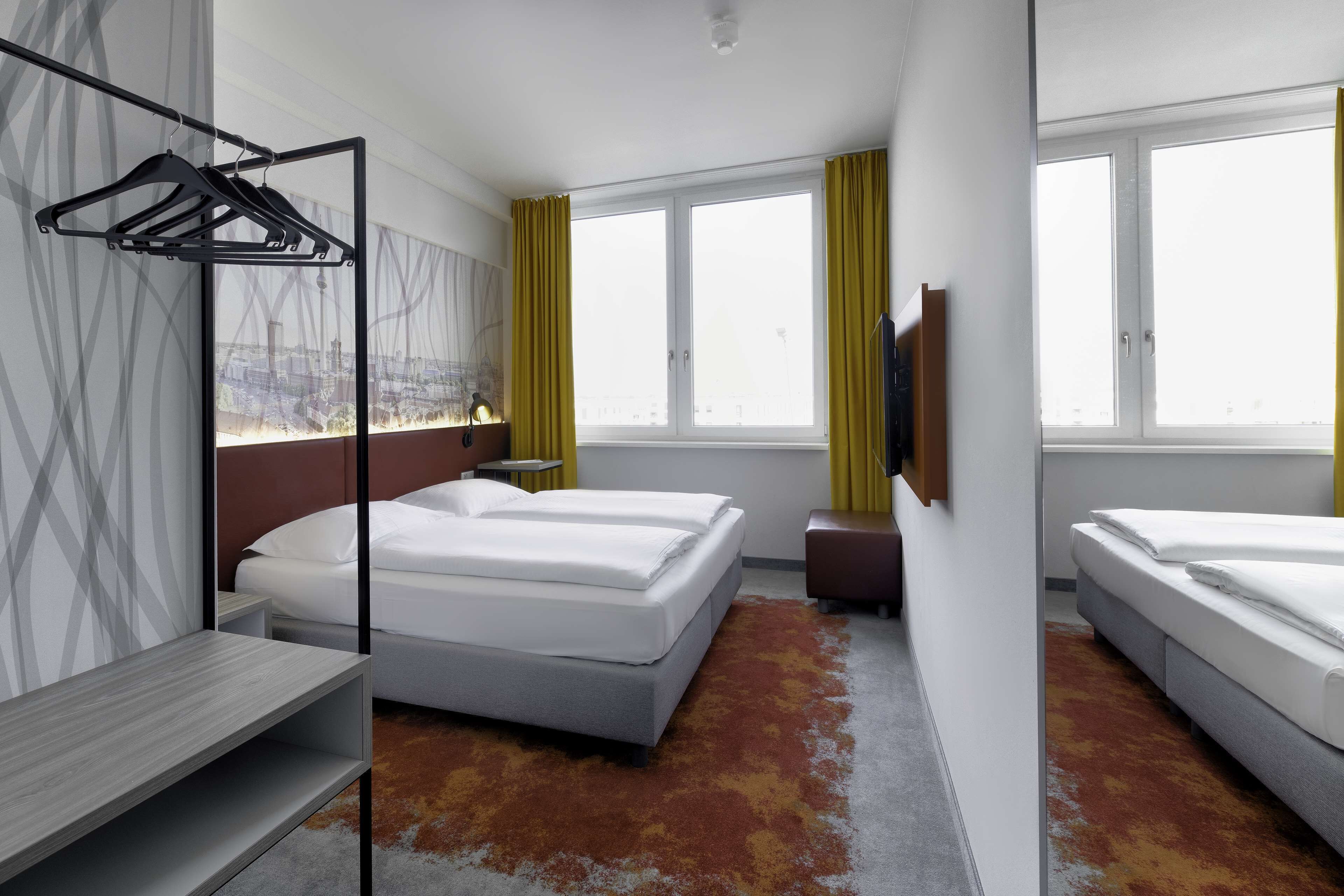 Habitación de hotel con dos camas, un escritorio, perchero y un amplio ventanal con cortinas.