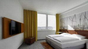 Habitación de hotel con cama, televisión montada en la pared, ventana con cortinas y vista a la ciudad.