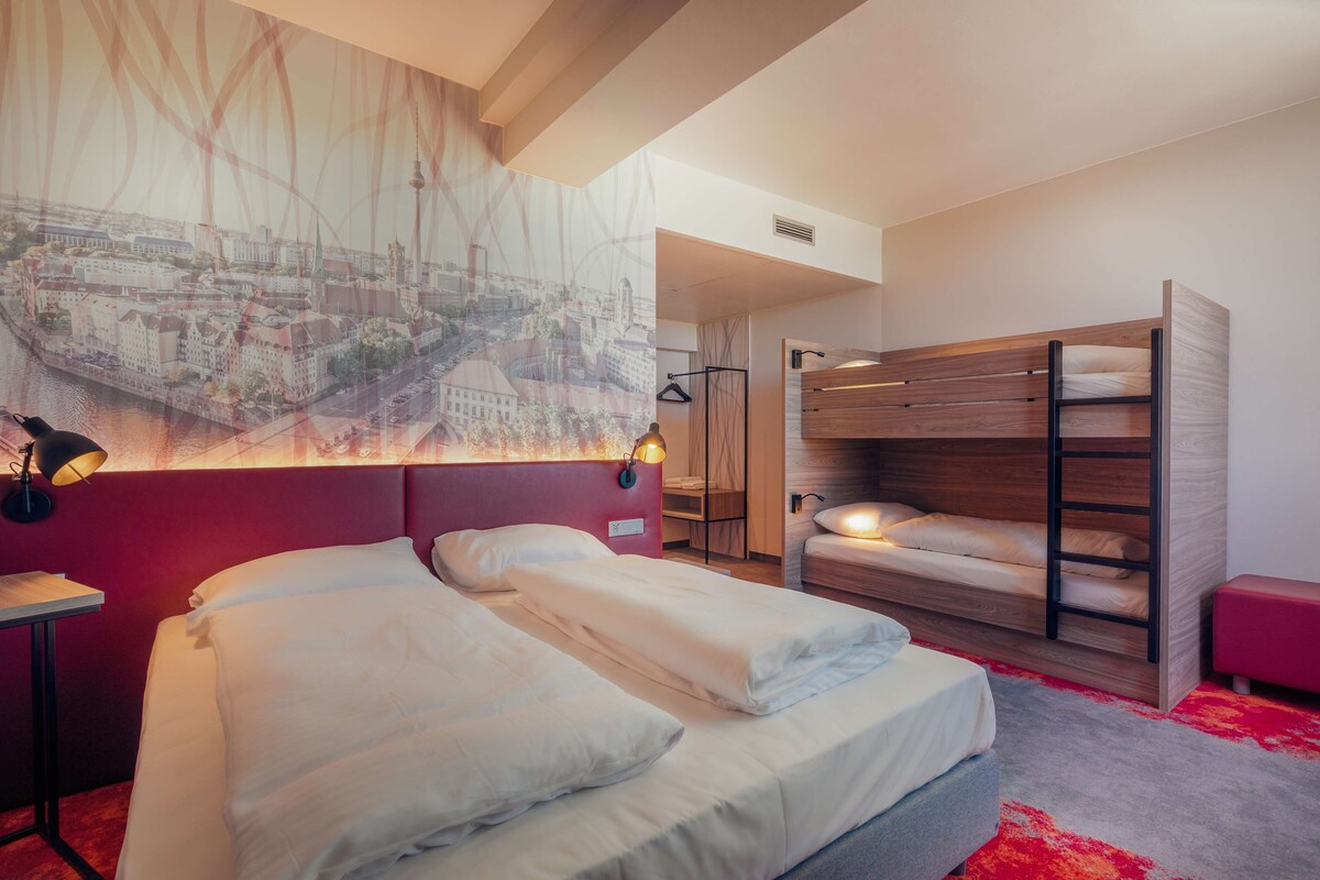 Habitació d'hotel amb un llit gran, una lliterà i escriptori amb làmpada, i mural amb vista a la ciutat a la paret.