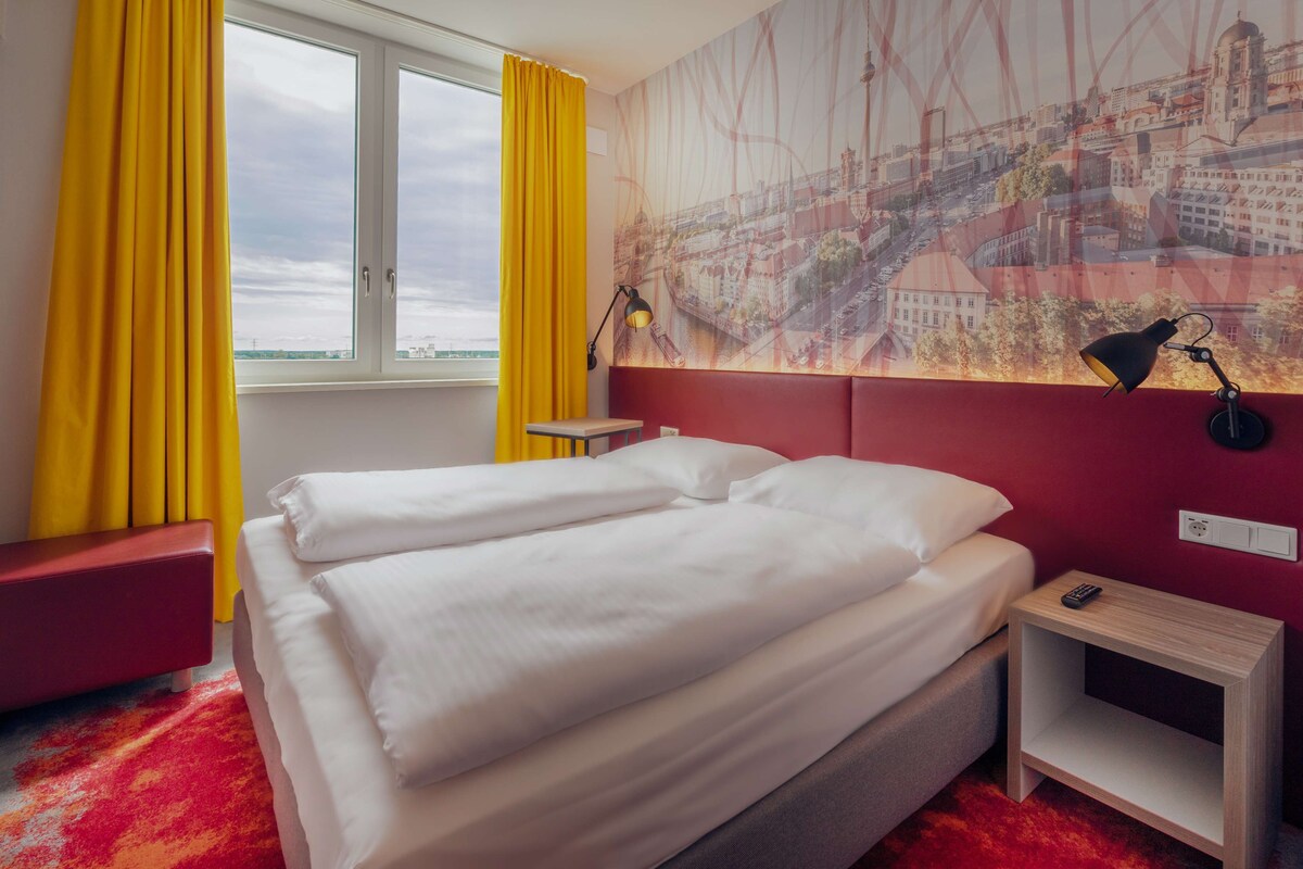 Habitació d'hotel amb un llit, una tauleta de nit, una panxa vermella, una finestra amb cortines grogues i un mural de paisatge urbà a la paret.