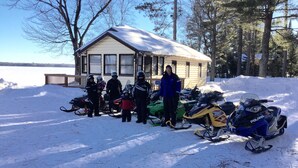 A cabin with a snow-covered roof, surrounded by snowmobiles and people in winter gear. - Pet-Friendly Cabin On The Lake With Fireplace (St. Germain)