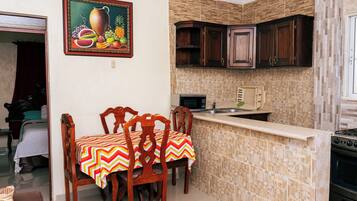 A dining area with a table set for four, wooden chairs, and a colorful tablecloth. A kitchen with stone backsplash and dark wooden cabinets is visible.