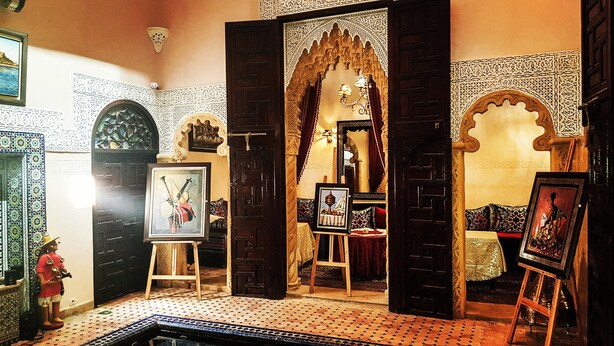 A room with a Moroccan-style interior, featuring a wooden door with intricate carvings, a tiled floor, and various framed artworks. - Riad La Porte Du Bouregreg (Salé)