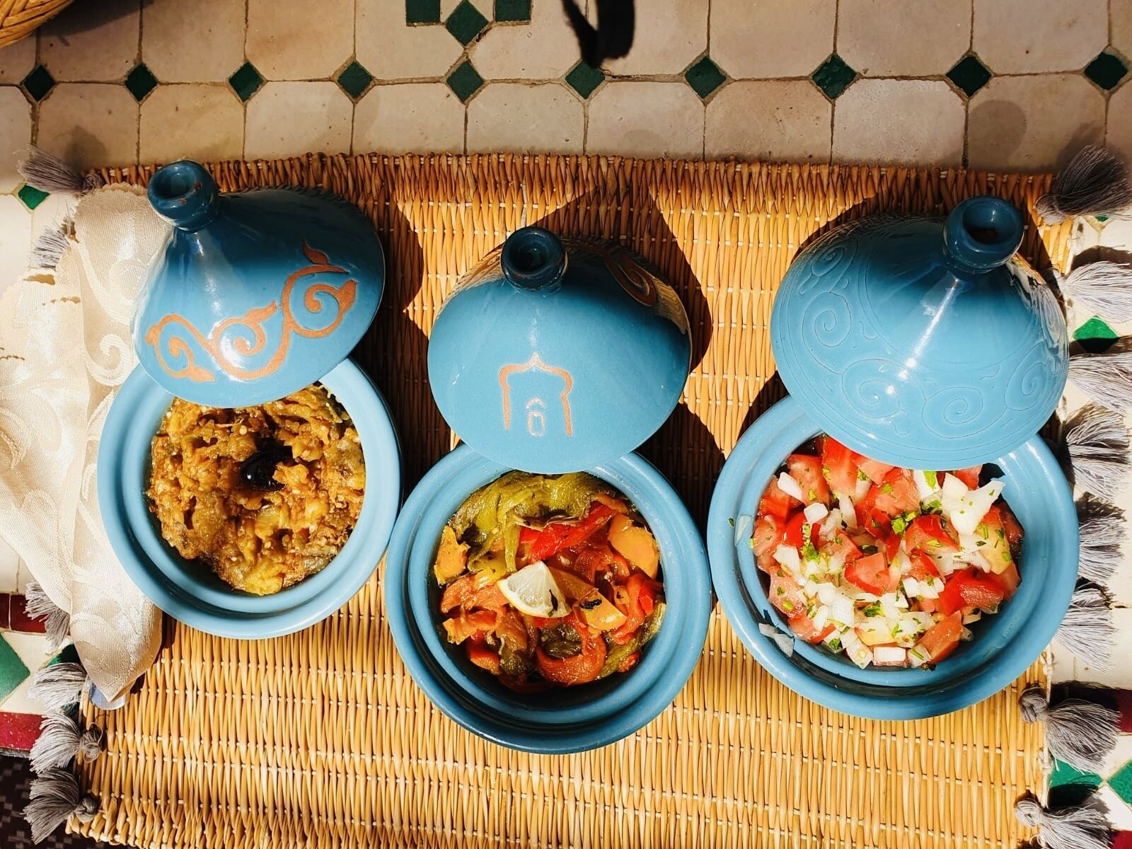 Three blue ceramic dishes with lids, each containing different food items, placed on a woven mat.