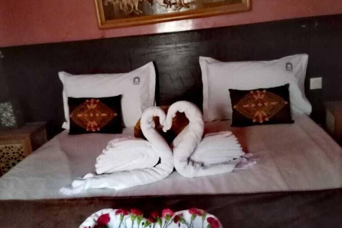 Una cama bien tendida con toallas dispuestas en forma de corazón, una almohada decorativa y un arreglo floral sobre la cama.