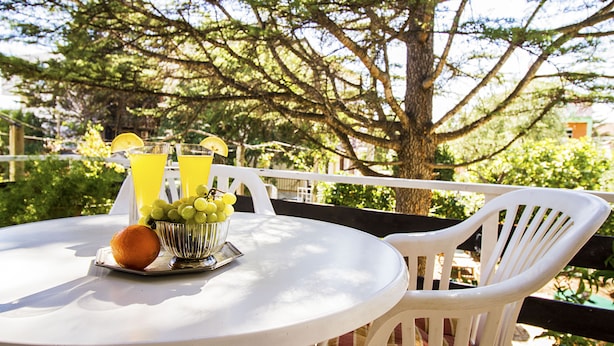 A round white table with a fruit bowl and two glasses of juice, surrounded by white chairs, set outdoors with trees in the background.