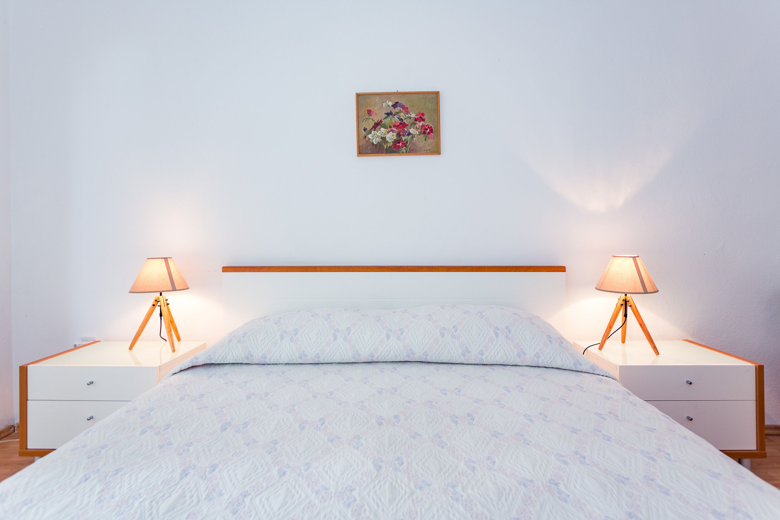 A neatly made bed with a patterned bedspread, two bedside tables with lamps, and a framed floral artwork on the wall.