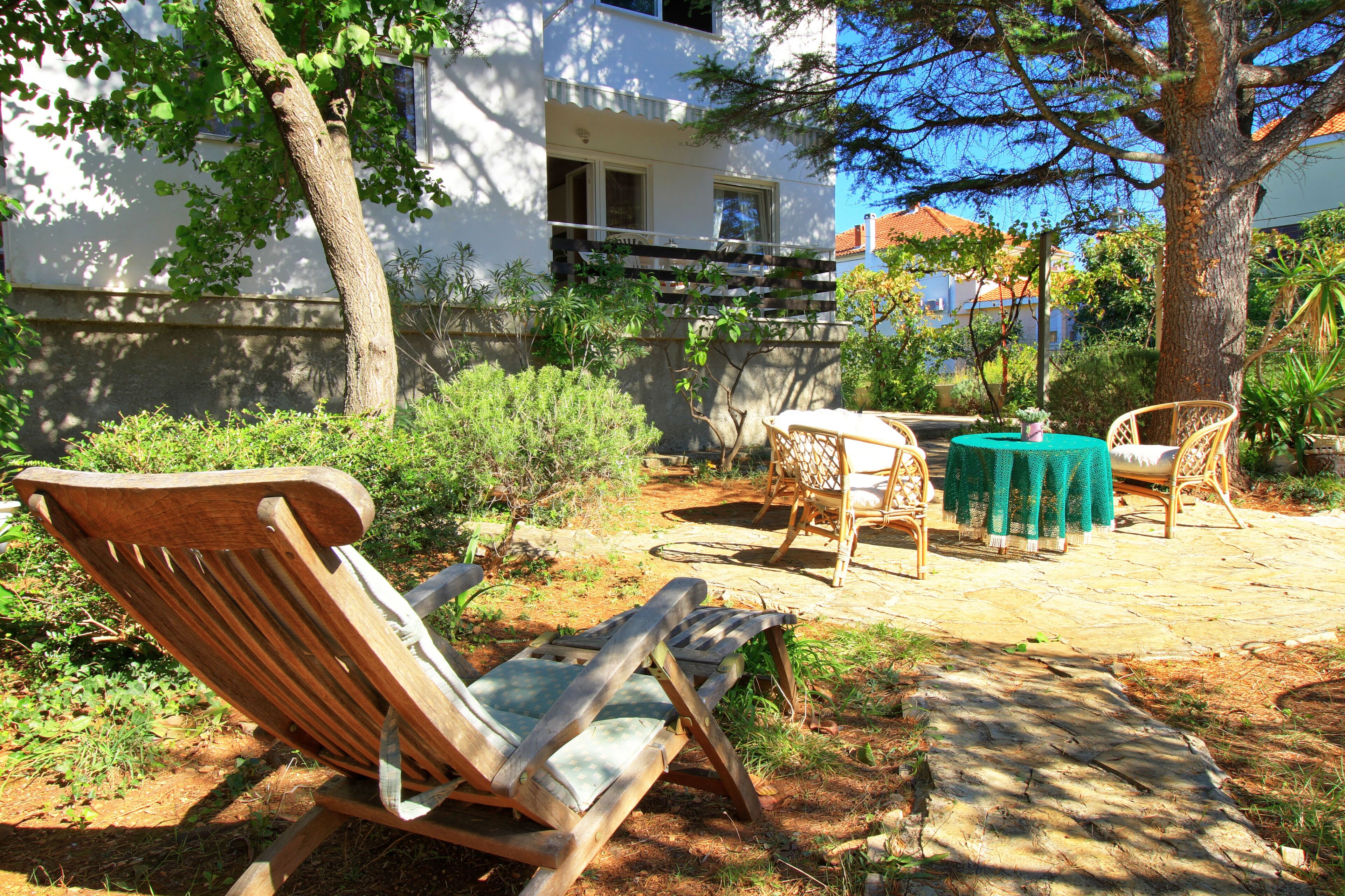 An outdoor seating area with wooden chairs and a table, surrounded by greenery and a building in the background.