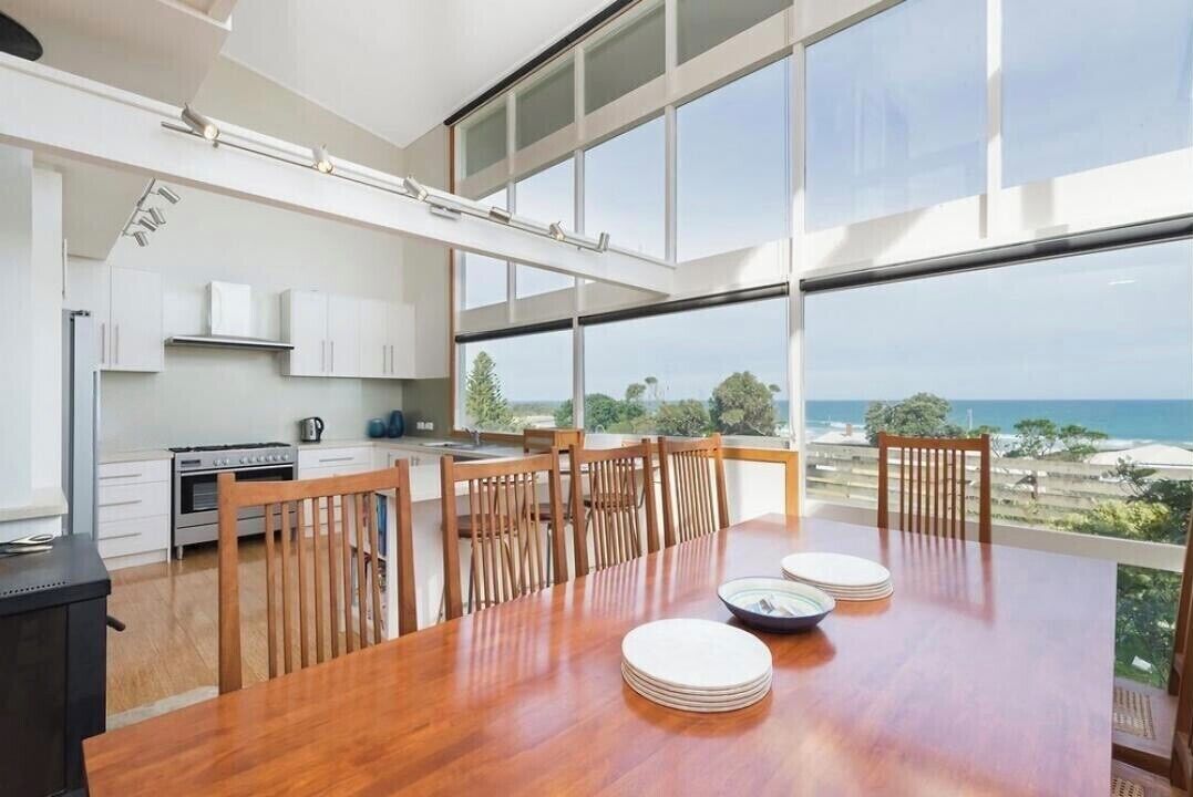 A modern kitchen with a large dining table, wooden chairs, and a view of the ocean.