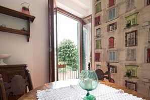 A dining table with a glass and lace tablecloth, wooden chairs, a shelf with a candle, and a view of a building with shutters.