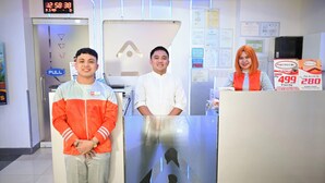 Three individuals standing in a hotel reception area with a digital clock displaying 12:58:38. - Astrotel Guadalupe (Makati)
