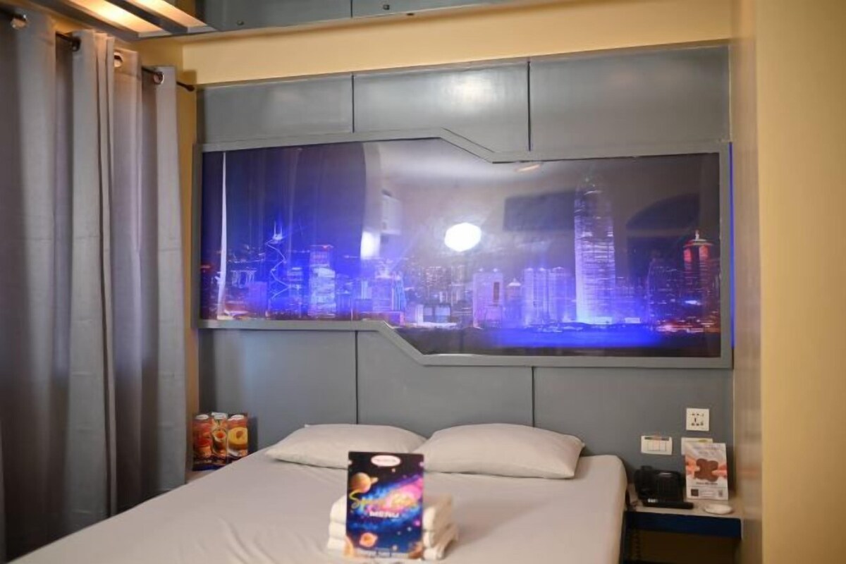 A hotel room with a bed, a nightstand, and a wall-mounted TV displaying a cityscape.
