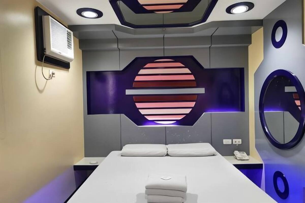A modern hotel room with a large bed, a built-in air conditioning unit, a wall-mounted mirror, and a unique geometric light fixture.