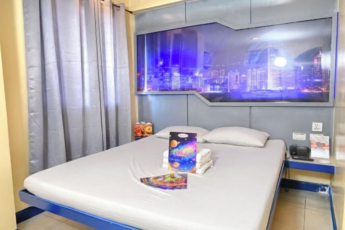 A hotel room with a bed, a cityscape view, curtains, and a nightstand with a phone and a book.