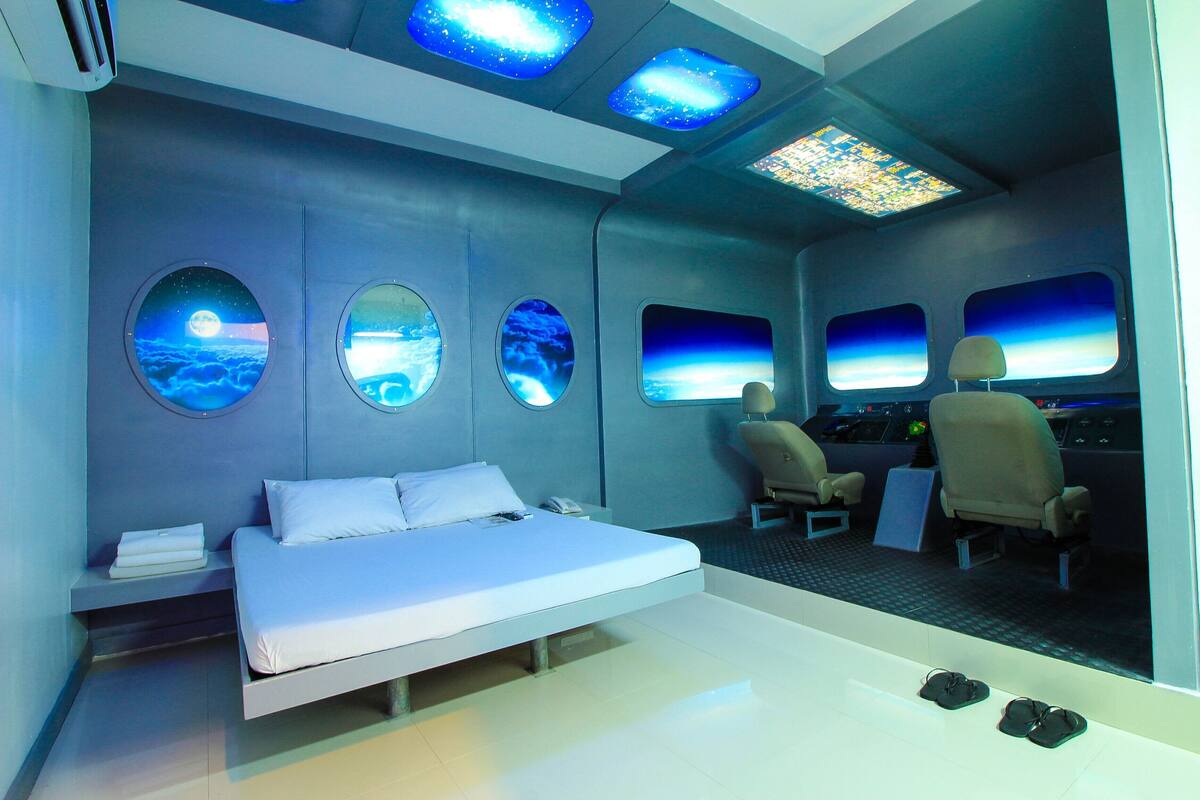 A modern room with a bed, two chairs, and three windows with views of the sky.