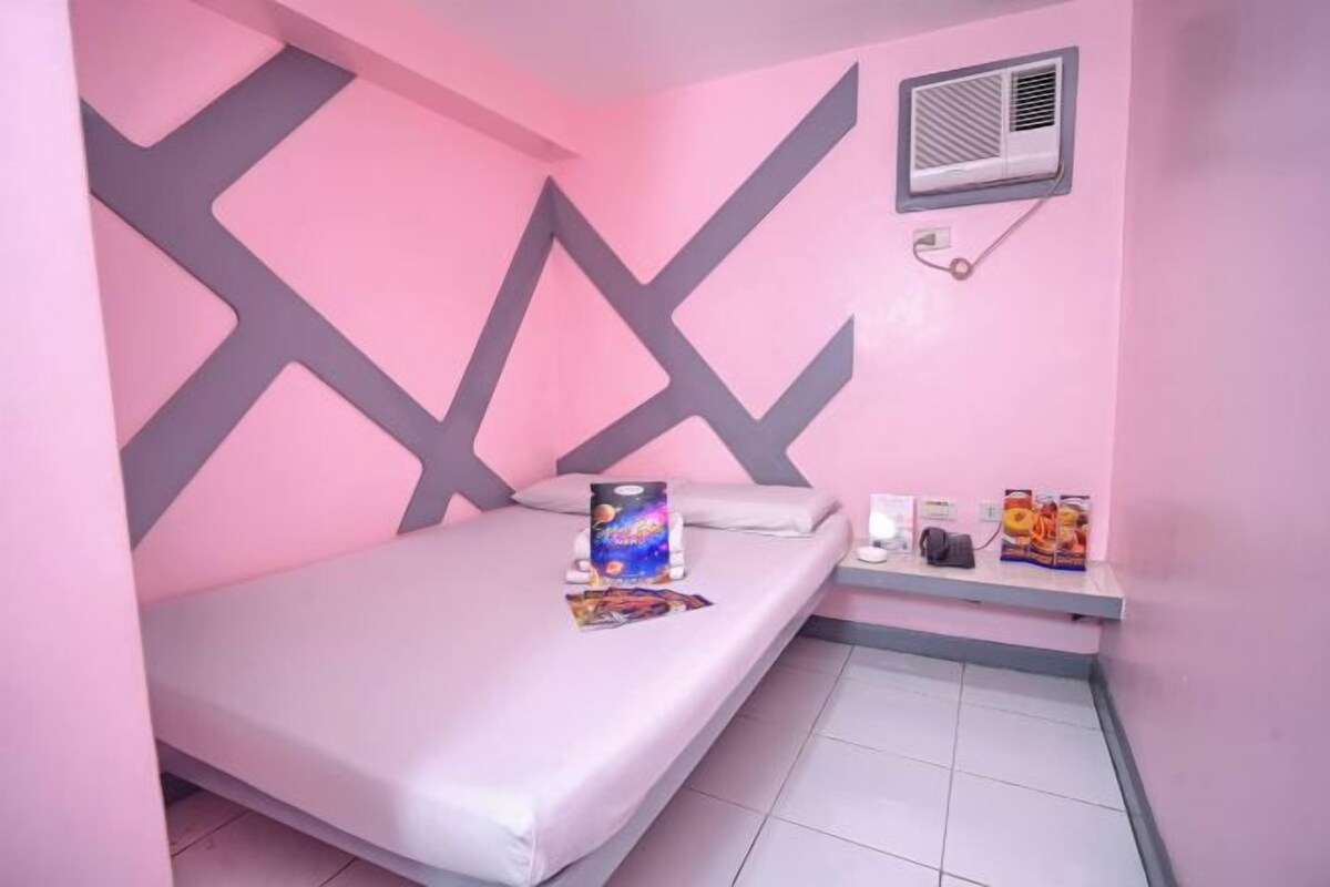 A small, pink-themed room with a bed, a bedside table, and a wall-mounted air conditioning unit.