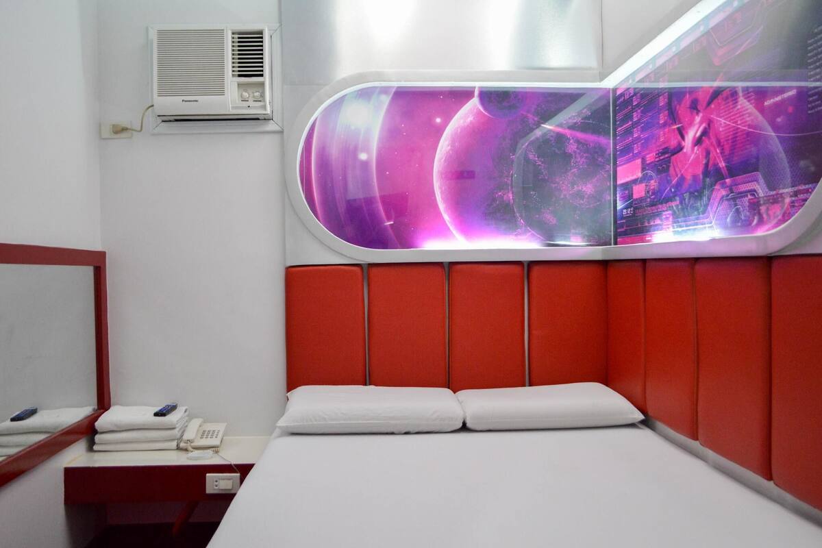 A hotel room with a modern design, featuring a bed with white linens, a red padded headboard, and a large window with a view of a space-themed digital display.
