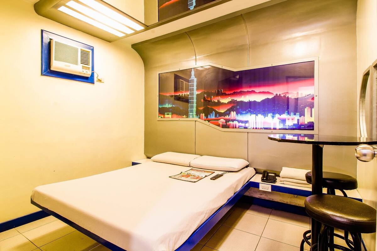 A hotel room with a bed, a small table with a chair, a wall-mounted air conditioner, and a cityscape artwork.