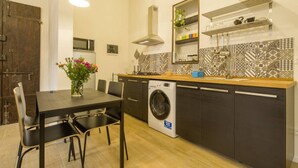 A compact kitchen with a washing machine, a dining table with chairs, a hanging light fixture, and a decorative mirror.