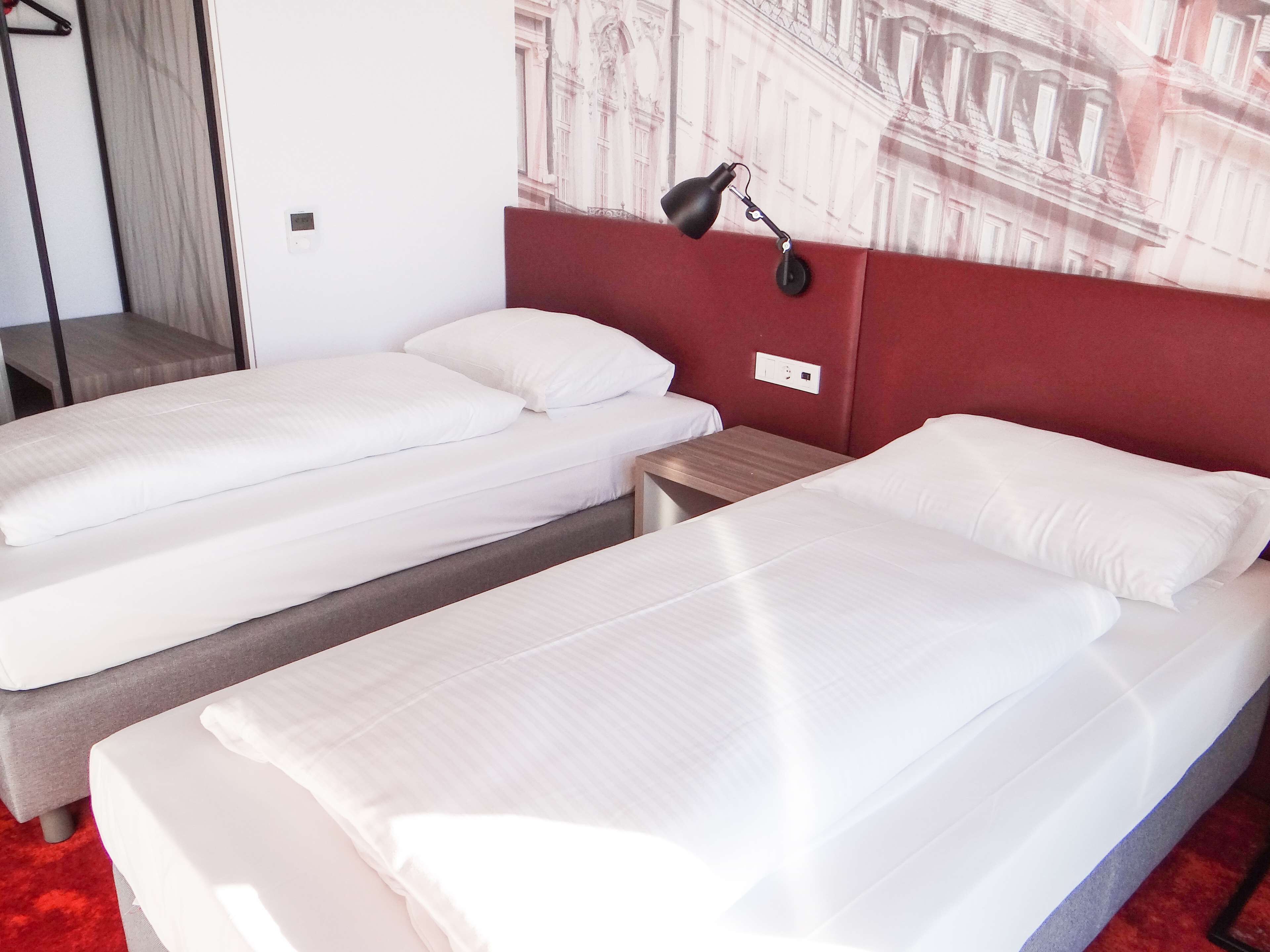 A hotel room with two single beds, a red headboard, a nightstand, a lamp, and a wall-mounted power outlet.
