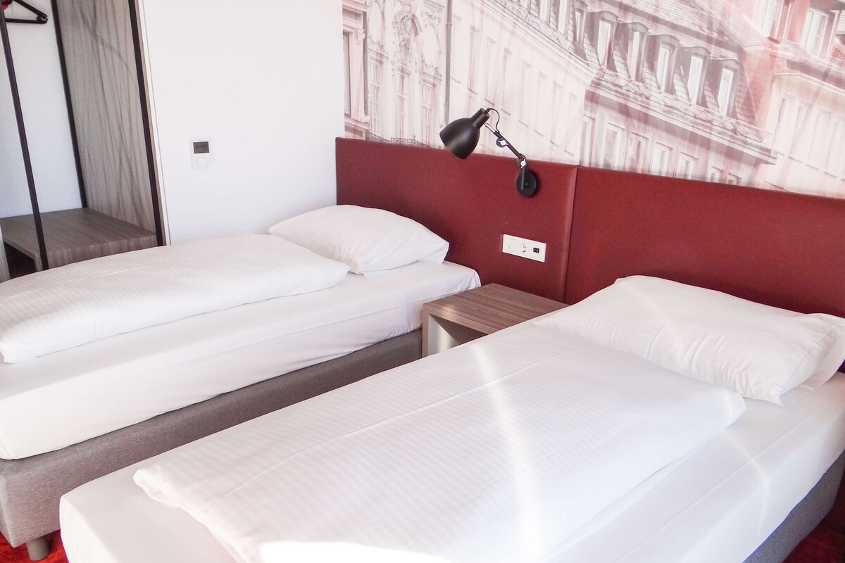 Une chambre d’hôtel avec deux lits simples, une tête de lit rouge, une table de chevet, une lampe et une prise de courant murale.