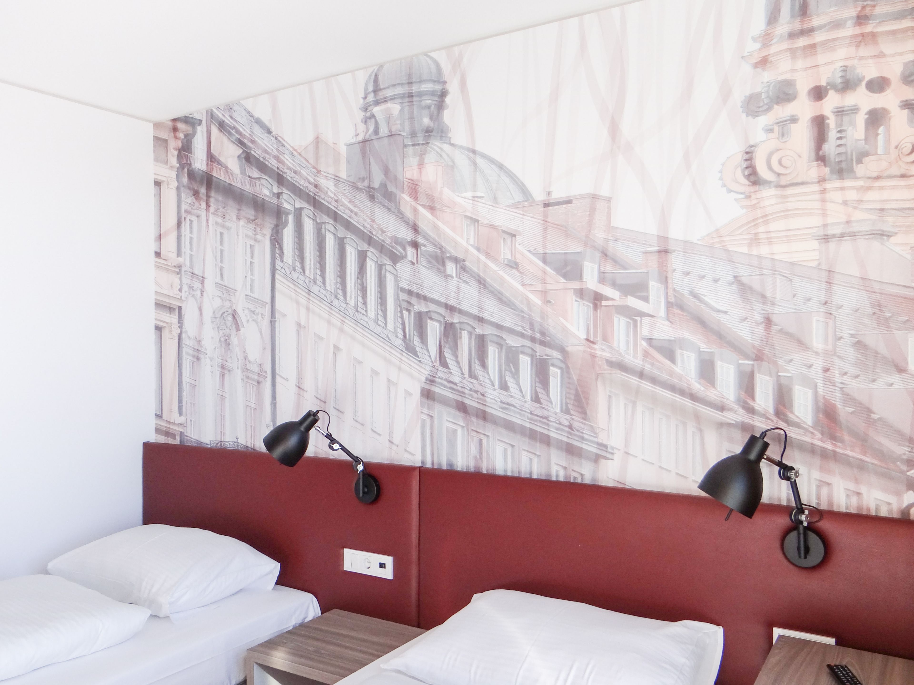 A hotel room with two beds, a red accent wall, and a large cityscape mural.