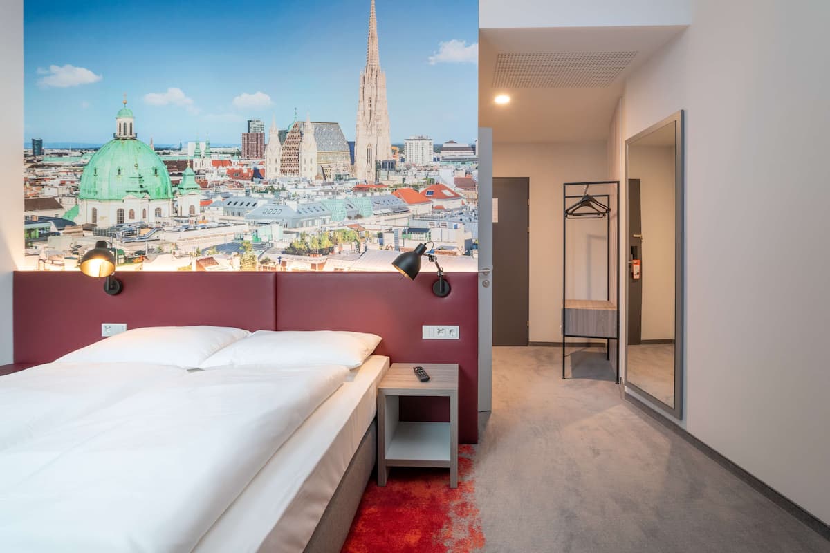 Una habitación de hotel con una cama, una mesita de noche, un espejo y un mural en la pared con un paisaje urbano.