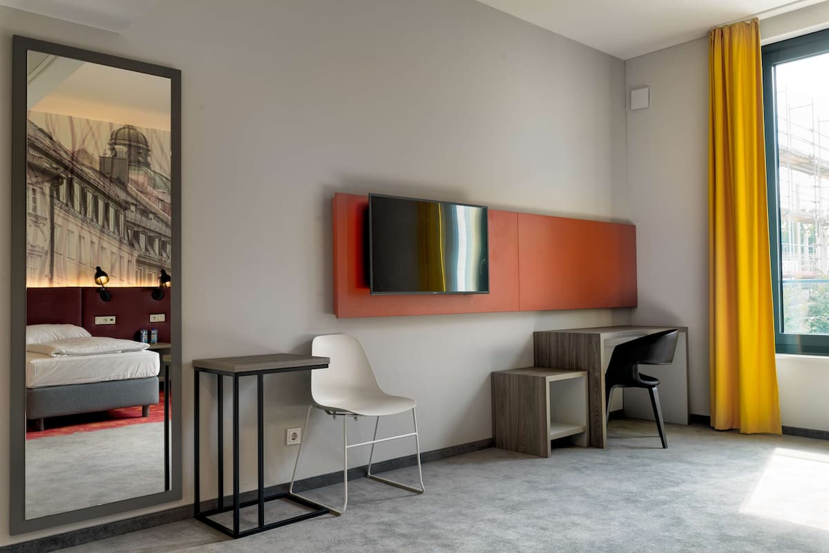 Una habitación de hotel moderna con cama, escritorio, silla, televisor y un amplio ventanal con cortinas.