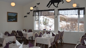 A dining area with tables set for guests, a painting on the wall, and windows with a view of a snowy landscape. - Huts in Víðidalur (Hvammstangi)