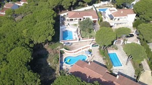 An aerial view of a residential property with multiple swimming pools, a large house with a tiled roof, and surrounding dense greenery.