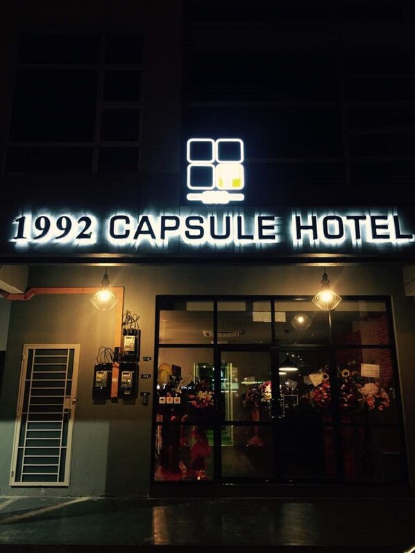 Exterior del Hotel Cápsula 1992, con un letrero de neón, grandes puertas de vidrio y decoraciones interiores a la vista.