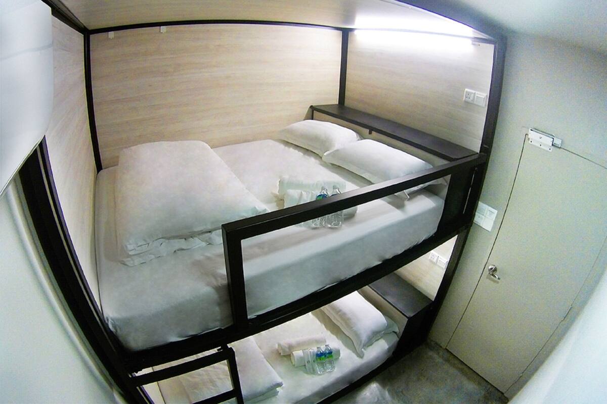 Una cama litera con ropa de cama blanca y estructura de madera. En la litera inferior, hay una mesita con una botella y una taza. Se ve un armario blanco a la derecha.