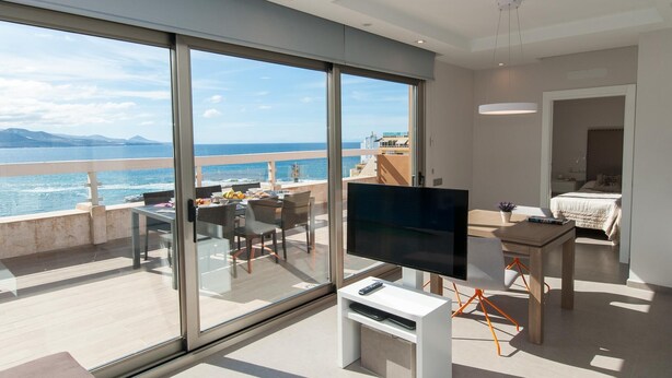 Un salon moderne doté d’un grand téléviseur, d’un coin repas avec une table et des chaises, et d’un balcon offrant une vue sur la mer.