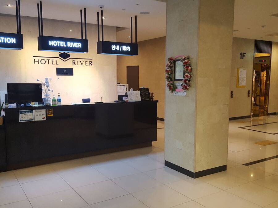 A hotel reception desk with a sign that reads "HOTEL RIVER" and "INFORMATION" above it. There is a digital screen, a plant, and a small table with items on it.