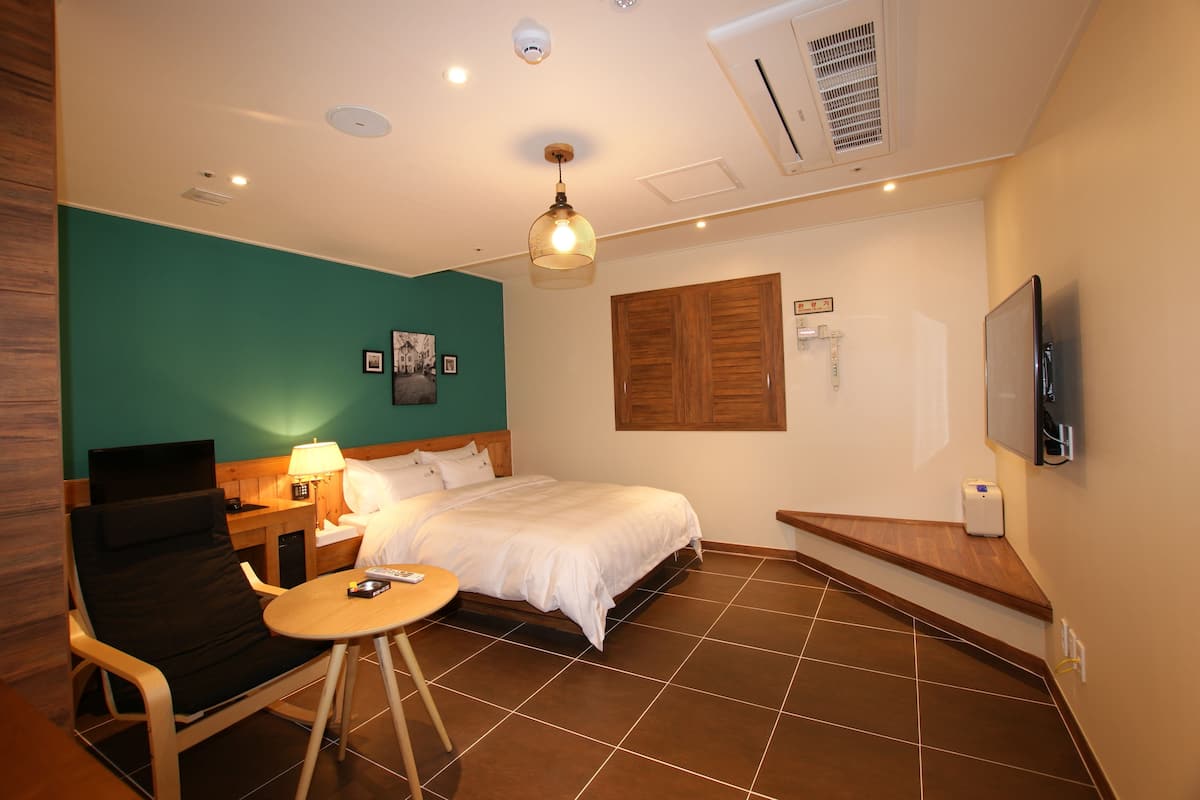 A hotel room with a bed, a chair, a small table, a lamp, a TV, and a wall-mounted air conditioner.