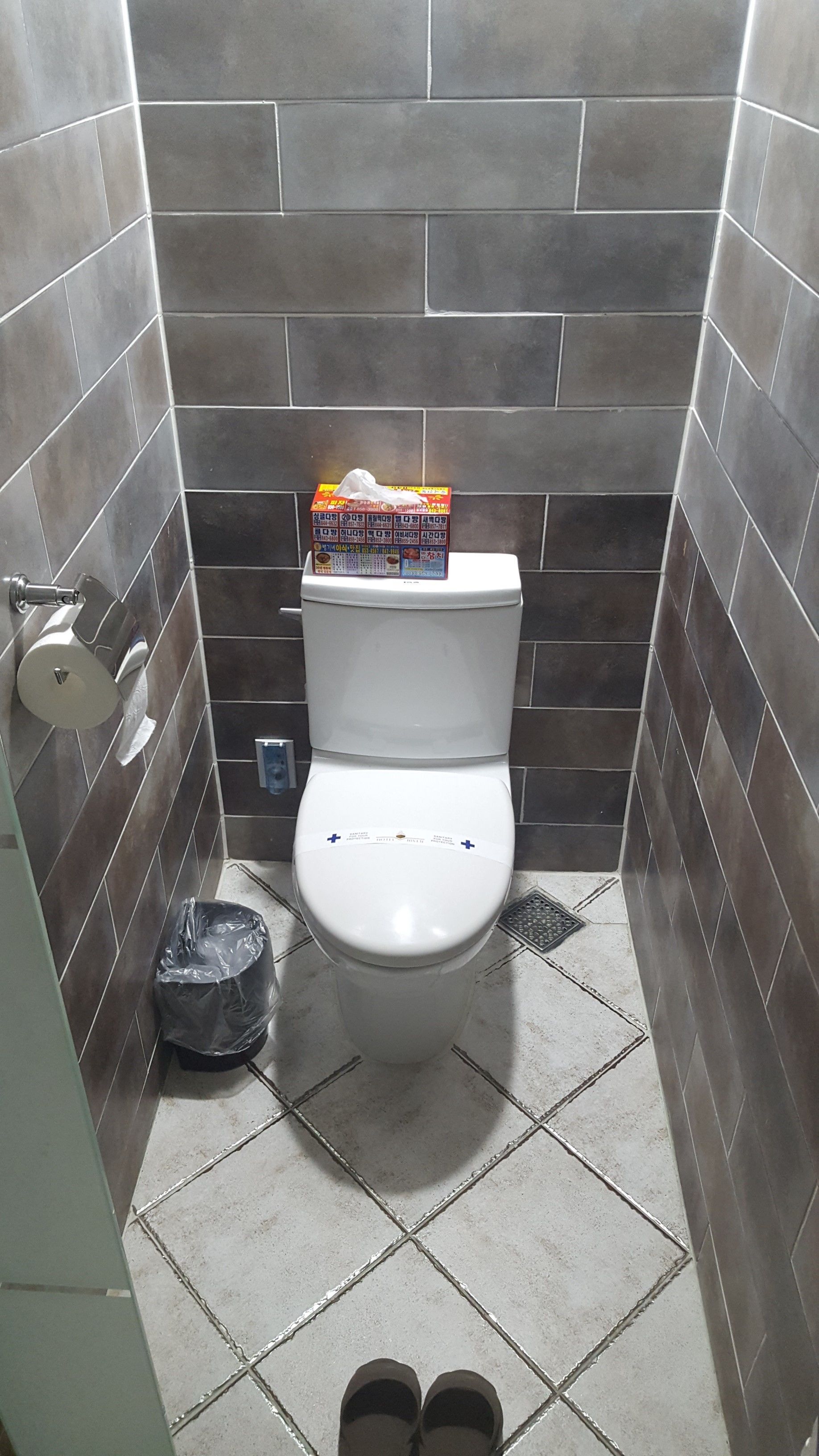 A small, tiled bathroom with a toilet, a trash bin, and a toilet paper holder.