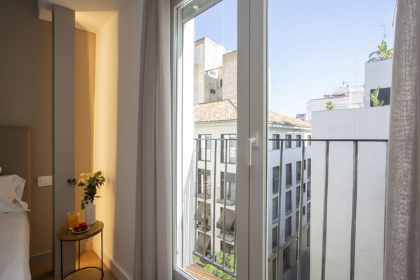 A hotel room with a large window, a bedside table with a plant, and a view of adjacent buildings.