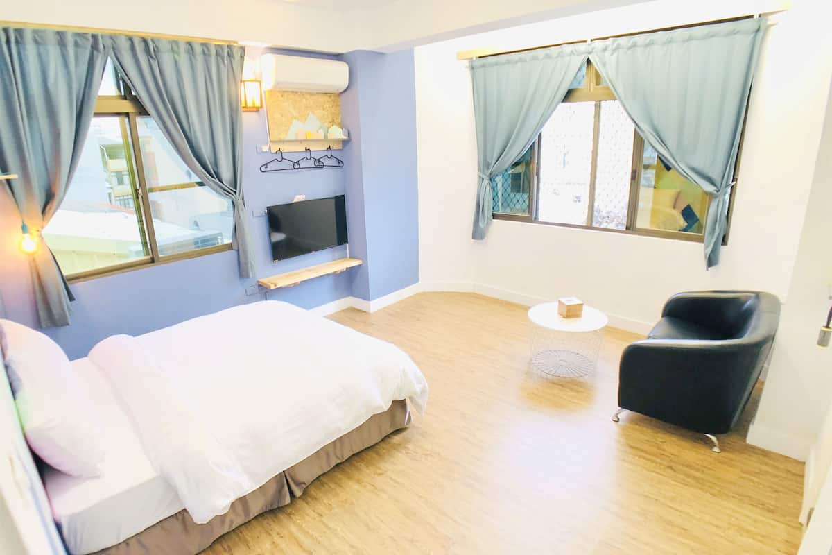 A hotel room with a bed, a TV mounted on the wall, a small table, a chair, and a window with curtains.