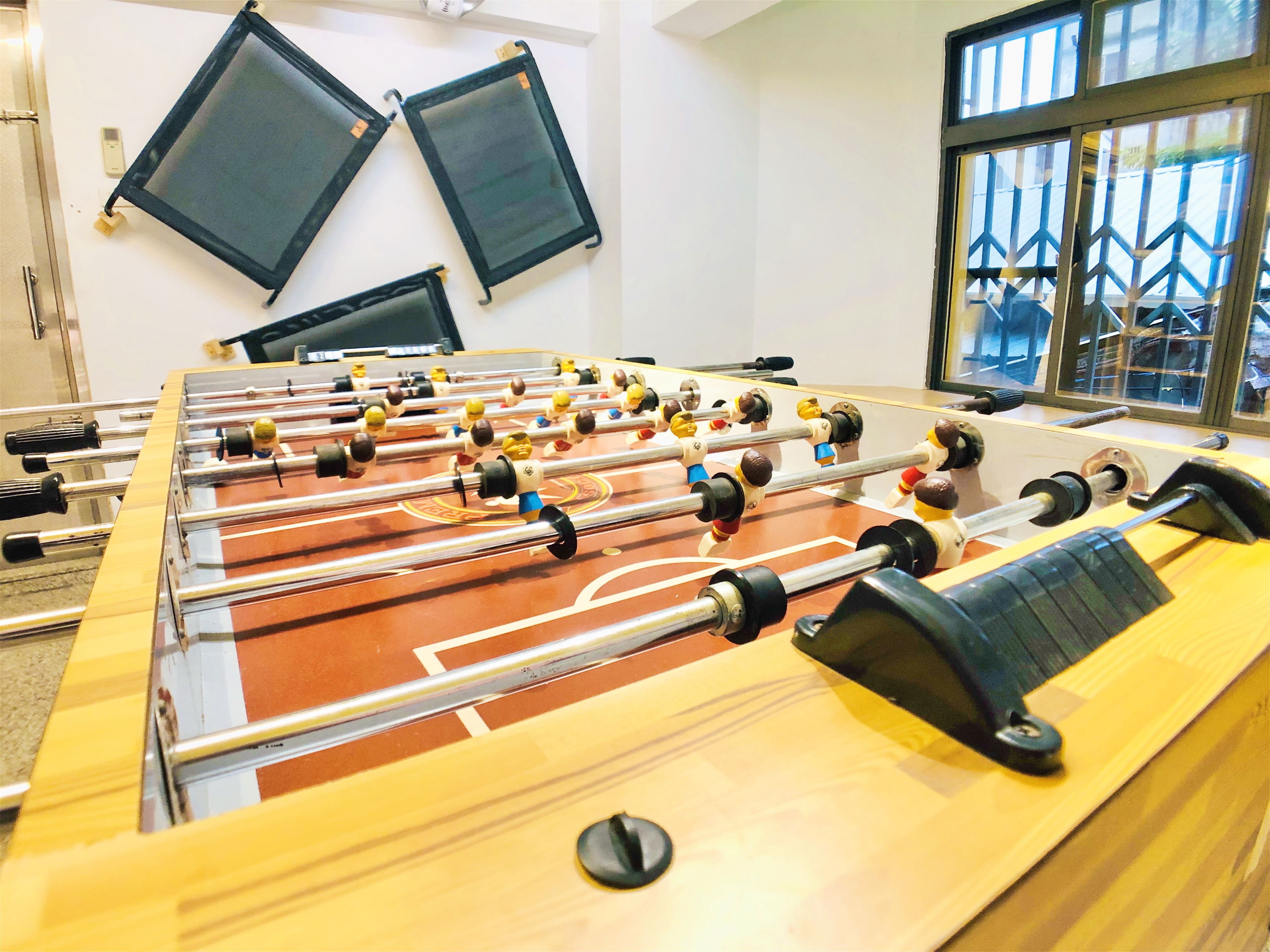 A foosball table in a room with two monitors on the wall and a window with a view of a building.