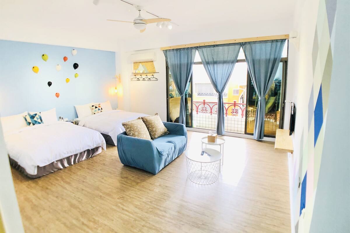 A modern hotel room with two beds, a blue sofa, a small round table, and large windows with curtains.