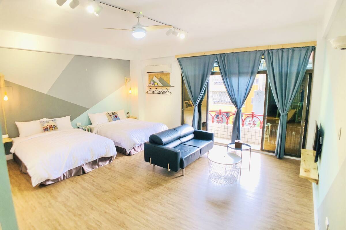 A hotel room with two beds, a sofa, a small table, and a balcony with a railing.